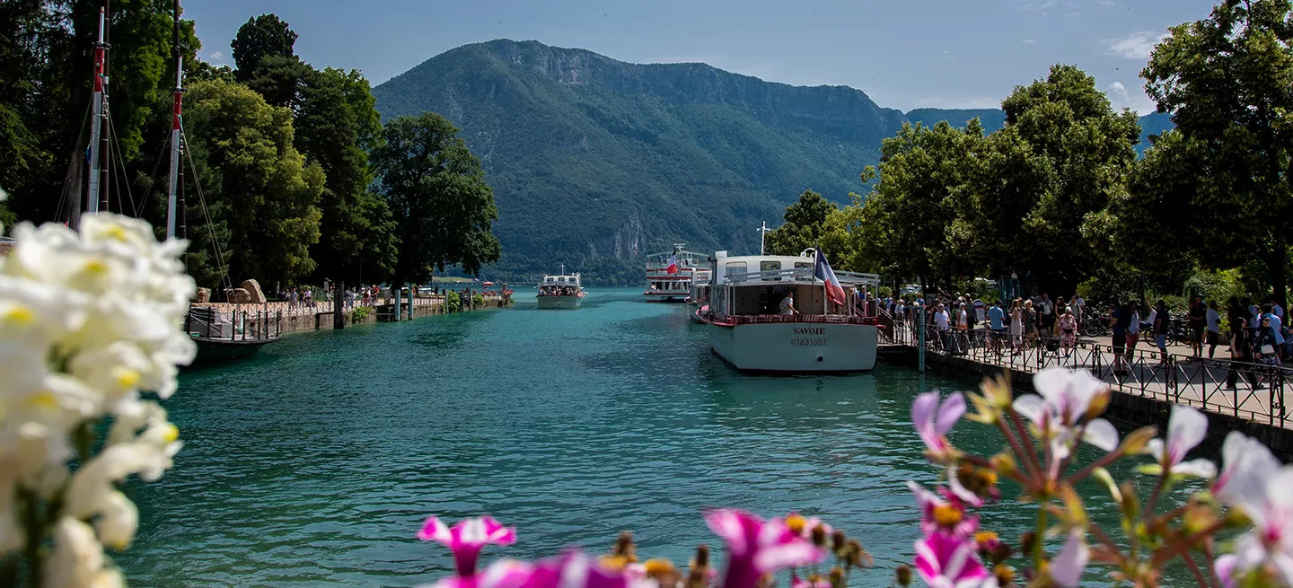 A view of turquoise Lake Annecy with white boats, French flags, green trees, hazy mountains, blurred flowers.