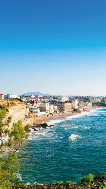 A view of the coastal city of Biarritz from a cliff on a sunny day, waves crashing against its beach.